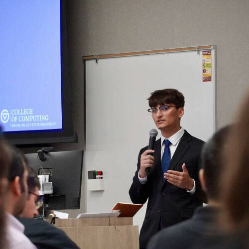 A student in a dark suit, blue tie, and glasses speaks into a microphone during a presentation at Grand Valley State University’s College of Computing. The presentation slide beside him displays the college’s logo and title while audience members l...